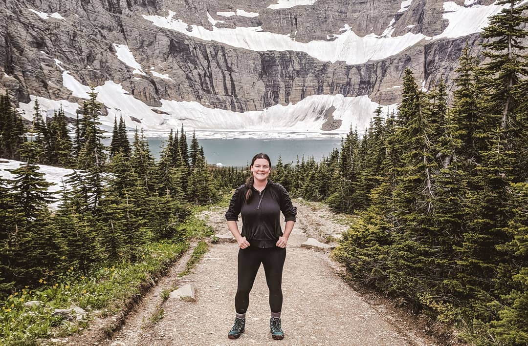 A hiker stands in all-black clothing in front of the bright turquoise waters of Iceberg Lake in Glacier National Park