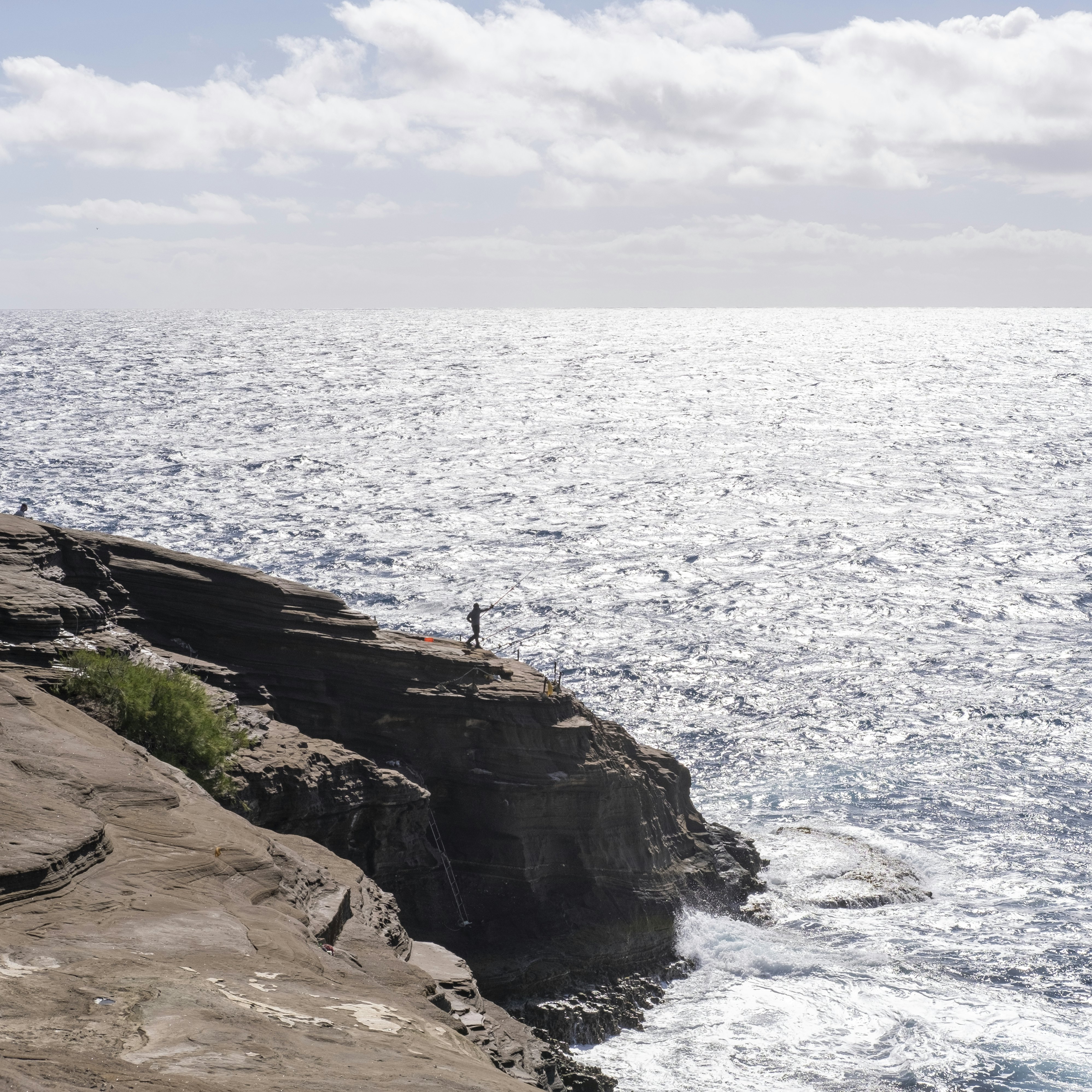 There is a fisherman about to cast his line in the water over a tall cliff. Spitting Caves Hawaii is a hidden gem of East Oahu.