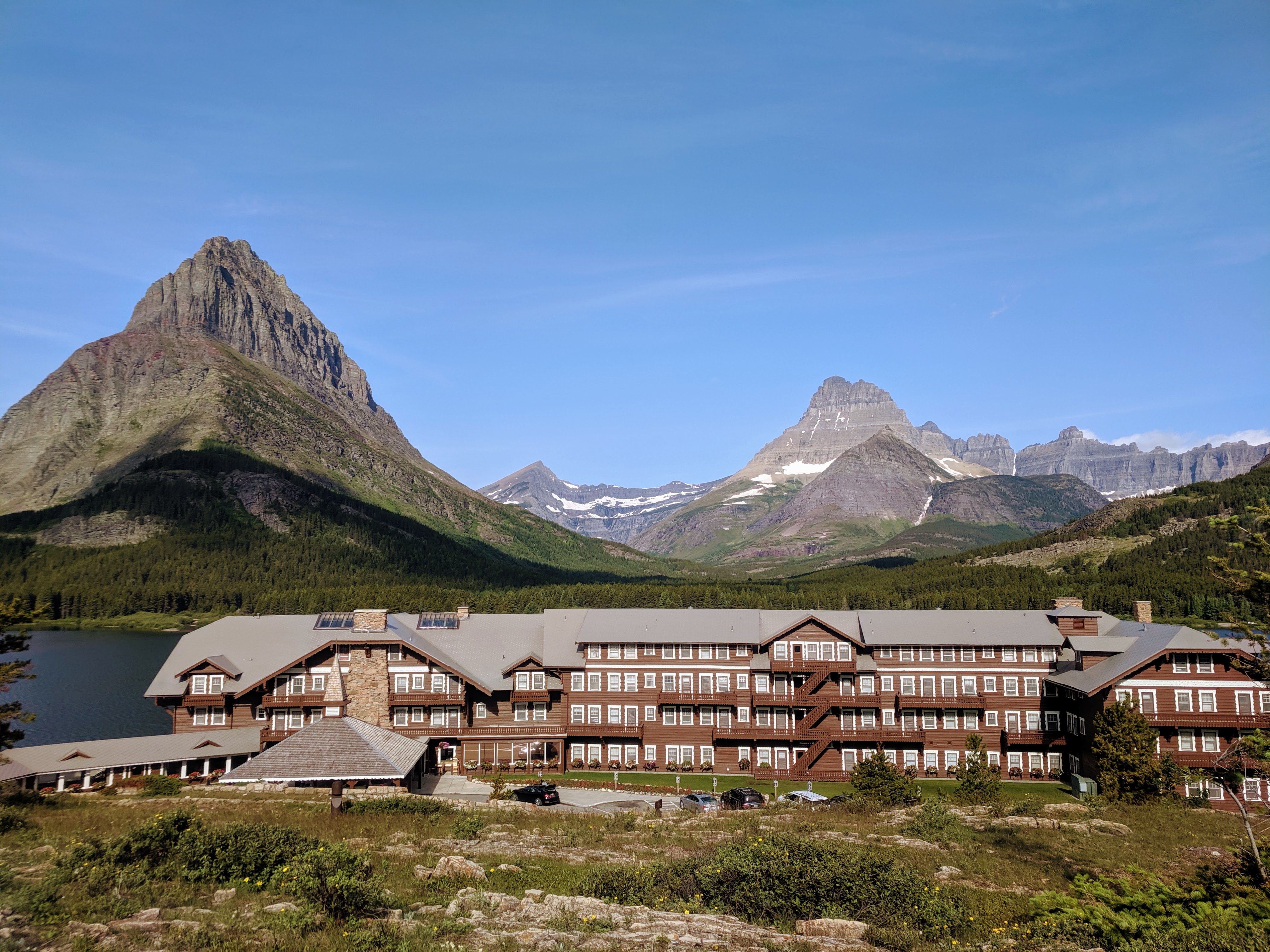 A classic alpine lodge sits by a bright blue lake under towering, jagged grey mountain peaks