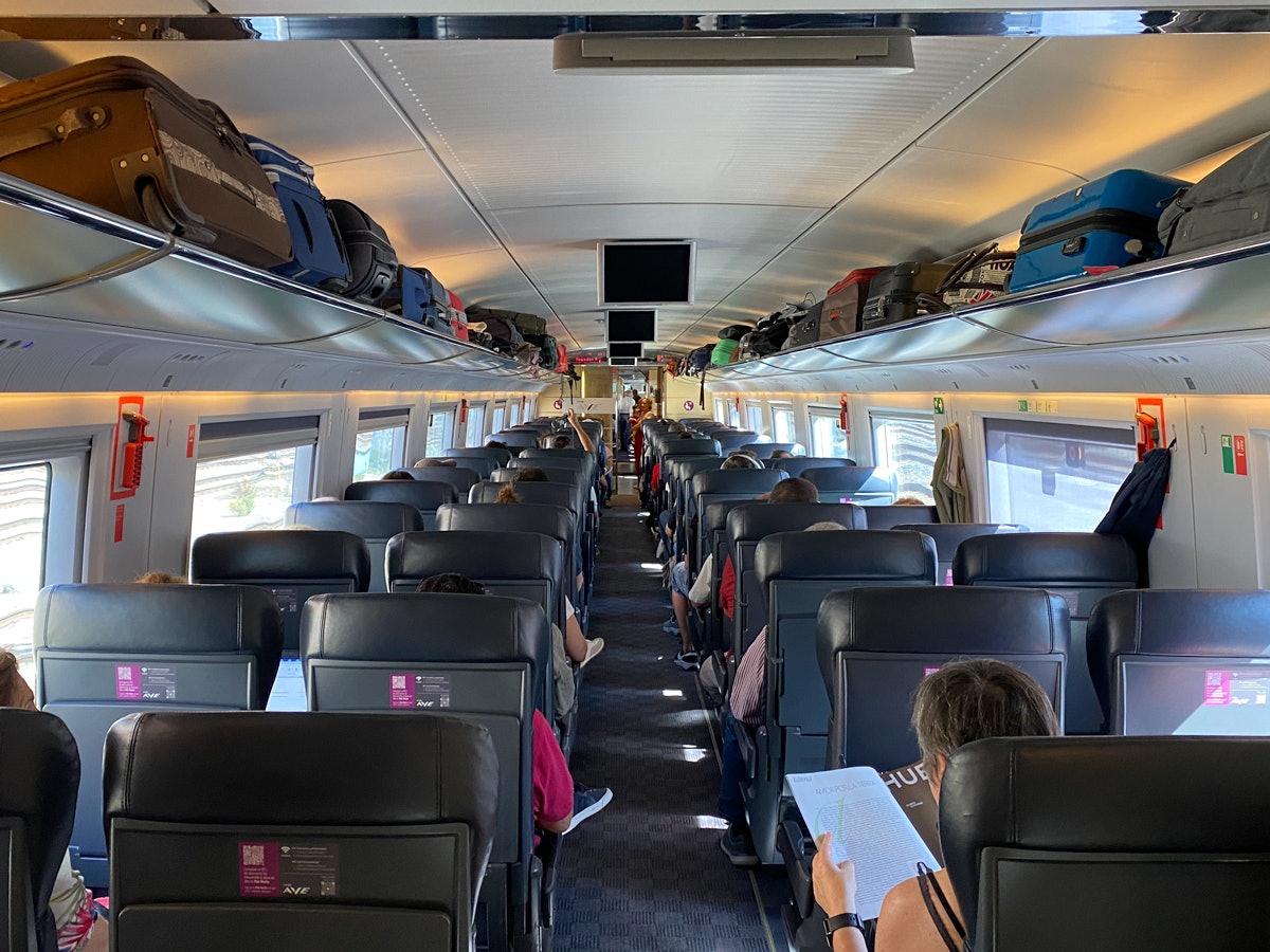 Passengers sat in seats in a 2-by-2 formation in a modern train carriage. Luggage is in the racks above them.