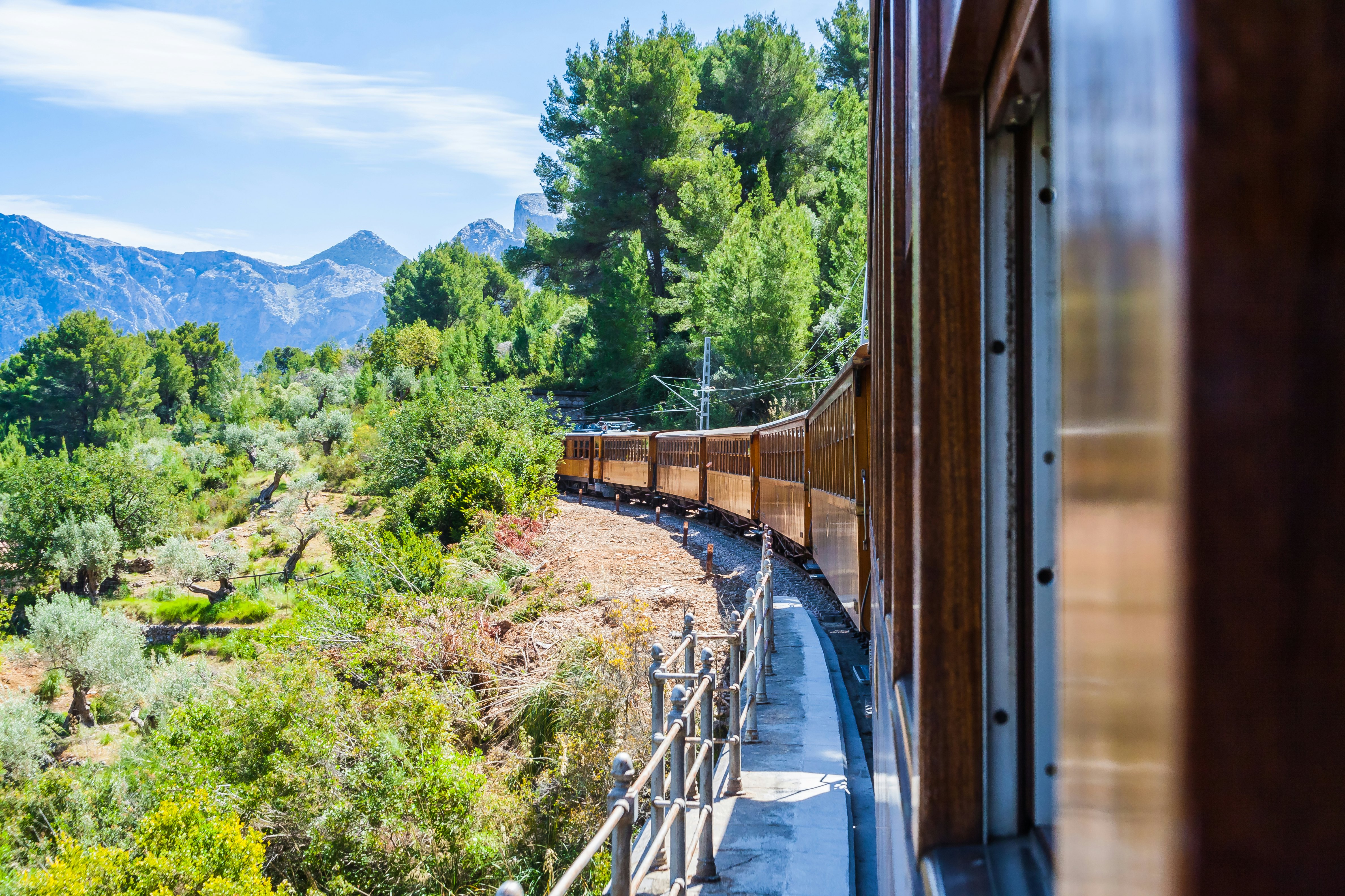 A vintage train with wooden carriages trundles through countryside and mountains on a sunny day.